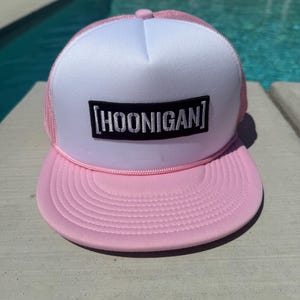 May include: Pink and white trucker hat with the word "HOONIGAN" in a black rectangle. The hat has a pink brim and a white front panel. The mesh on the sides is pink.