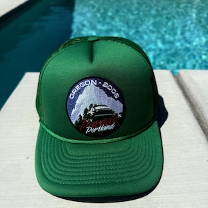 May include: A green trucker hat with a circular patch featuring the text "Oregon - 2006 Parade Portland". The patch has a mountain and vehicle graphic in blue, white, and red. The hat is sitting on a white surface with a pool in the background.