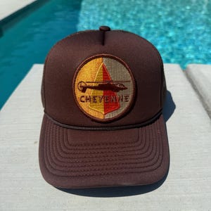 May include: A brown trucker hat with a mesh back and a curved brim. The front features a circular patch with a helicopter design and the word "CHEYENNE" in red lettering. The hat has a dark brown rope detail and a button on top.