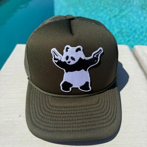 May include: Olive green trucker hat with a black mesh back and a white and black panda bear patch holding two guns. The hat has a curved brim and a green rope detail.
