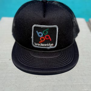 Könnte beinhalten: Schwarze Trucker-Mütze mit gebogenem Schirm und Mesh-Rückseite. Die Mütze hat einen quadratischen Aufnäher mit dem Text "breckenridge" und den Zahlen "69" in Blau, Grün und Rot.