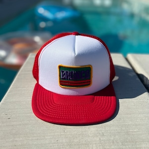 May include: A red and white trucker hat with a green, purple, and yellow patch that says "CROWNED" on it.