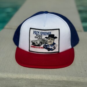 May include: A red, white, and blue trucker hat with a mesh back. The front panel features a patch with the text "Palm Springs Road Races" and an illustration of a vintage car and a man in a hat.