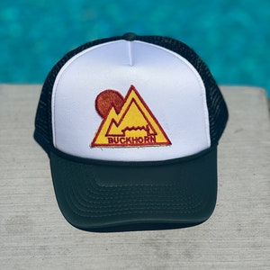 Könnte beinhalten: Eine weiße und schwarze Trucker-Kappe mit einem gelben und roten Bergmotiv-Patch, auf dem "BUCKHORN" steht.