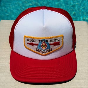May include: Red and white trucker hat with a red mesh back and a curved brim. The front features a patch with text and a logo. The hat has a classic, casual style.