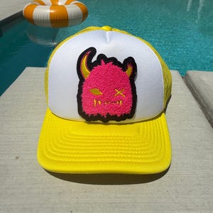 May include: A yellow and white trucker hat featuring a pink monster patch. The hat has a yellow peak and mesh back, with a white front panel. The monster patch is pink with yellow horns and a black outline. The hat is on a concrete surface.