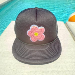 Puede incluir: Gorra de camionero negra con un parche floral. La flor es rosa con centro amarillo y contorno azul claro. La gorra tiene una visera curva y una parte trasera de malla.