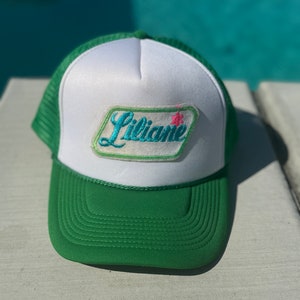 May include: A white and green trucker hat with a green mesh back. The hat has a patch on the front that says "Liliane" in a teal blue cursive font with a pink star.