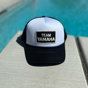 May include: A white and black trucker hat with a black patch that says "TEAM YAMAHA" in white letters. The hat is sitting on a wooden surface near a swimming pool.