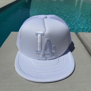 Puede incluir: Gorra blanca de camionero con las letras "LA" bordadas en la parte delantera. La gorra tiene una parte trasera de malla y una visera curva. La gorra está sobre una superficie de hormigón.