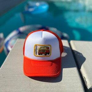 May include: A white and orange trucker hat with a patch featuring a cartoon yellow and red Chevy van with the word "CHEVY" above it.