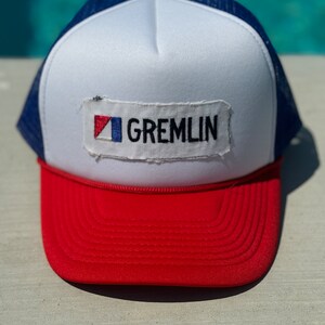 May include: A white, red and blue trucker hat with a patch that says "GREMLIN" in black letters. The patch has a small red, white and blue flag design in the top left corner.