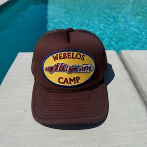 Puede incluir: Gorra de camionero marrón con parte trasera de malla y visera curva. La parte delantera de la gorra presenta un parche con el texto "WEBELOS CANYON BALL RUN 2002 CAMP" en amarillo y azul.