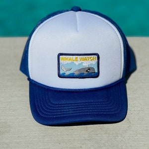 Könnte beinhalten: Blau-weiße Trucker-Cap mit einem Aufnäher "WHALE WATCH". Der Aufnäher zeigt einen Wal, der aus dem Wasser springt. Die Cap hat einen blauen Schirm und eine weiße Vorderseite. Sie besteht aus Mesh und Stoff.