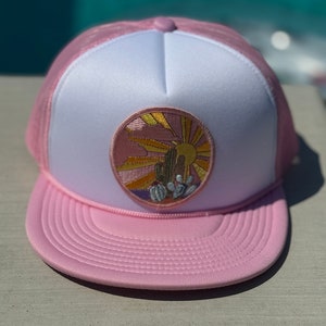 May include: A pink and white trucker hat with a pink and yellow embroidered patch of a cactus and a sun. The patch is on the front of the hat.