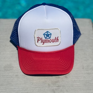 May include: A white and red trucker hat with a blue mesh back. The hat has a patch on the front that says "Plymouth" with a blue star logo.