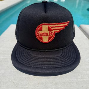 May include: Black trucker hat with a curved brim and mesh back. The hat features a circular patch with the words "Santa Fe" and a red and gold wing design. The hat is sitting on a light gray surface.