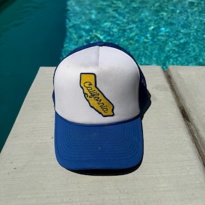 May include: A blue and white trucker hat with a blue brim and a white front panel. The front panel features a yellow outline of the state of California with the word "California" in a black font.