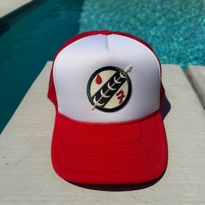 May include: A red and white trucker hat with a curved brim. The front panel is white with a circular emblem featuring a spear, blood drop, and the number 7. The hat has a red brim and mesh back.