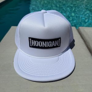 May include: White trucker hat with a curved peak and mesh back. The hat features the word "HOONIGAN" in black block letters on a rectangular patch. A white rope detail accents the peak.