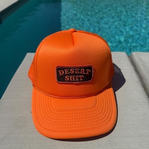 May include: Bright orange trucker hat with a mesh back and a curved brim. The hat features a rectangular patch with the words "DESERT SHIT" in a bold, serif font. The hat is sitting on a light-colored surface with a pool in the background.