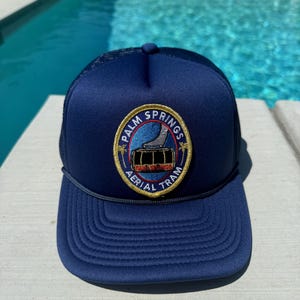 Op de afbeelding: Marineblauwe truckerpet met een Palm Springs Aerial Tram-patch. De patch toont een berglandschap en de woorden "Palm Springs Aerial Tram" in een cirkelvormig ontwerp. De pet heeft een mesh rug en een gebogen klep.