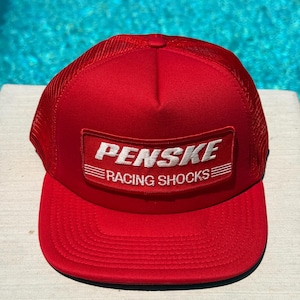 Peut inclure: Casquette de camionneur rouge vif avec un dos en filet et une visière incurvée. L'avant présente un écusson rectangulaire avec le texte blanc "PENSKE" au-dessus de "RACING SHOCKS". Un accessoire audacieux pour les amateurs de sport automobile.