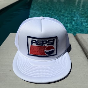 Pode incluir: Boné branco de caminhoneiro com aba curva e parte traseira em tela. A frente apresenta um patch retangular com a palavra "PEPSI" em azul marinho e o logotipo vermelho e azul da Pepsi.