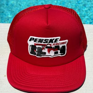 May include: A red trucker hat with a mesh back and a curved brim. The hat features a white and black Penske Racing logo patch on the front, depicting a race car. The hat is a vibrant red color.