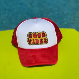 Puede incluir: Una gorra de camionero roja y blanca con un parche bordado amarillo y rojo que dice "GOOD VIBES".