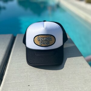 Puede incluir: Un gorro de camionero blanco y negro con un parche ovalado dorado que dice "Olympia Gold".