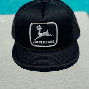 Op de afbeelding: Zwarte truckercap met een wit John Deere-logo met een hert in een vierkant. Het logo is geborduurd op de voorkant van de pet.