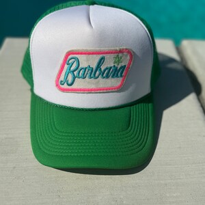 May include: A green and white trucker hat with a pink and blue patch that says "Barbara" in a cursive font.