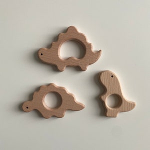 May include: Three wooden dinosaur teethers in natural wood tones. The teethers are shaped like a stegosaurus, a triceratops, and a tyrannosaurus rex.