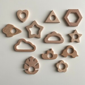 Könnte beinhalten: Ein Set mit 12 Holz-Beißringen in verschiedenen Formen, darunter ein Herz, ein Stern, eine Wolke, eine Rakete, eine Blume, ein Sechseck und ein Blatt. Die Beißringe sind alle in natürlicher Holzfarbe und haben eine glatte, polierte Oberfläche.
