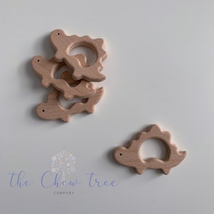 May include: Four natural wood dinosaur teethers stacked on top of each other. A fifth teether is lying on its side to the right of the stack. The teethers are shaped like dinosaurs with a ring in the middle. The teethers are made of natural wood and are unpainted. The text "The Chew Tree Company" is visible at the bottom of the image.