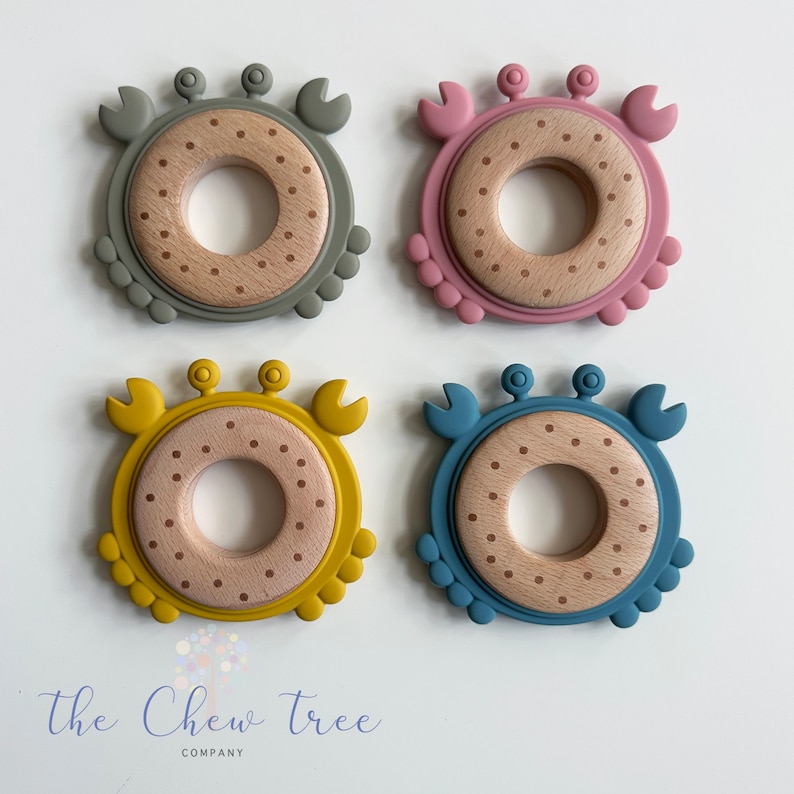 Puede incluir: Cuatro anillos de dentici&oacute;n de silicona con forma de cangrejo en diferentes colores: gris, rosa, amarillo y azul. Cada anillo tiene un centro de madera con lunares marrones.