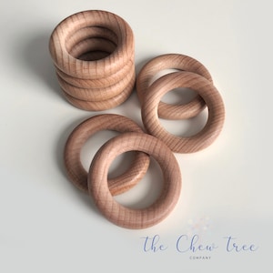 May include: A set of six natural wood teething rings. The rings are stacked on top of each other, with some rings laying flat on a white surface. The rings are smooth and have a natural wood finish.