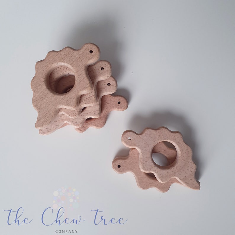 May include: A stack of four natural wooden teethers shaped like dinosaurs. The teethers have a hole in the middle and are designed for babies to chew on.