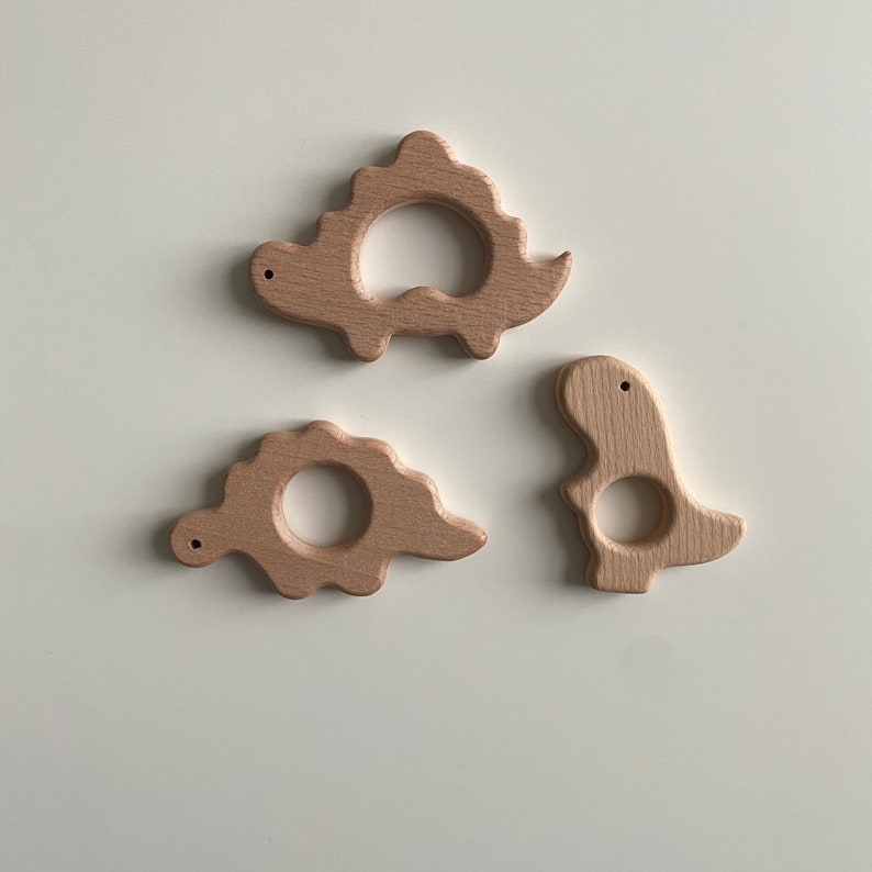 May include: Three wooden dinosaur teethers in natural wood tones. The teethers are shaped like a stegosaurus, a triceratops, and a tyrannosaurus rex.