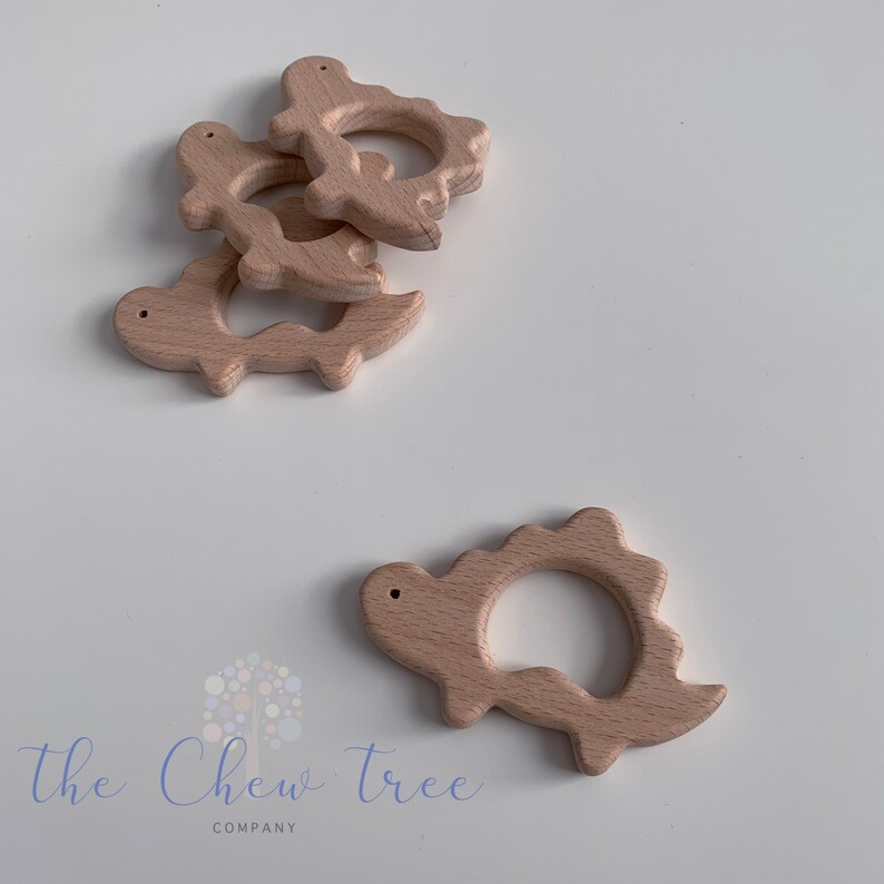 May include: Five natural wood dinosaur teethers. The teethers are shaped like dinosaurs with a ring in the middle. The teethers are a light brown color.