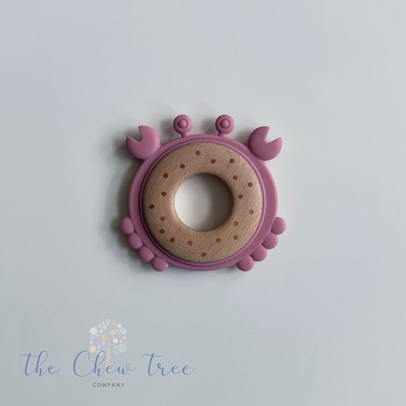 Puede incluir: Un anillo de dentici&oacute;n de silicona rosa con forma de cangrejo con un centro de madera marr&oacute;n. El anillo tiene un patr&oacute;n de puntos.