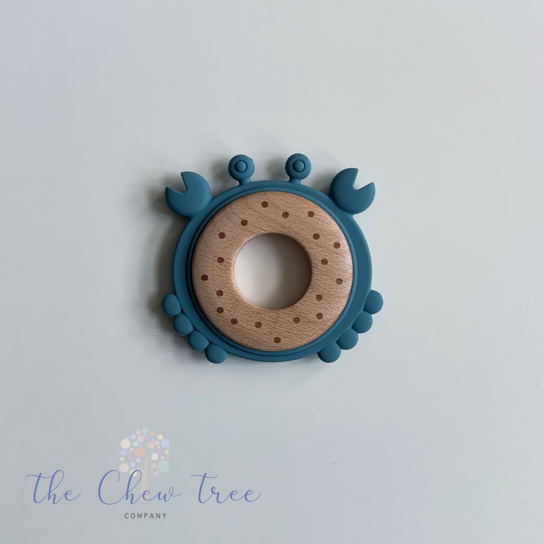 Puede incluir: Un anillo de dentici&oacute;n de silicona azul con forma de cangrejo con un centro de madera marr&oacute;n. El centro tiene forma de anillo y est&aacute; decorado con puntos marrones.