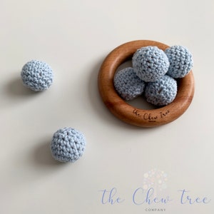 May include: A set of six light blue crocheted balls for babies to chew on. The balls are arranged in a wooden ring with the text "The Chew Tree Company" engraved on the ring.