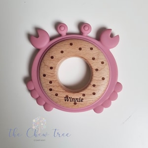 Puede incluir: Anillo de dentici&oacute;n de silicona rosa en forma de cangrejo con un anillo central de madera. El anillo de madera tiene lunares marrones y el nombre "Winnie" grabado en &eacute;l.