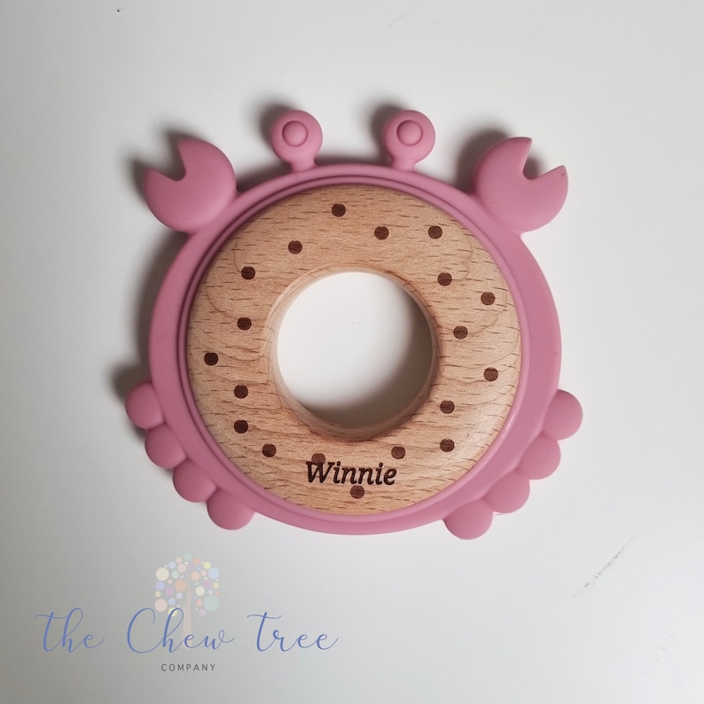 Puede incluir: Anillo de dentici&oacute;n de silicona rosa en forma de cangrejo con un anillo central de madera. El anillo de madera tiene lunares marrones y el nombre "Winnie" grabado en &eacute;l.