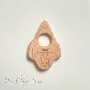 May include: A wooden rocket-shaped teether with a hole in the middle for a baby to hold. The teether is engraved with the name "Yusuf" and a star.