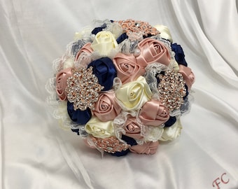Puede incluir: Un ramo de novia hecho de rosas de satén marfil, rosa empolvado y azul marino con adornos de encaje y pedrería.
