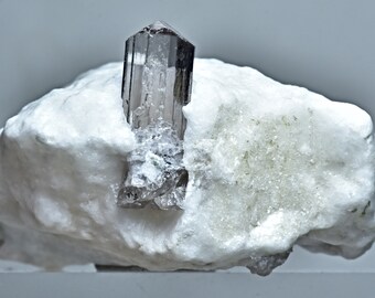 May include: A clear, smoky brown crystal with a pointed top, embedded in a white rock. The crystal is about 2 inches tall.