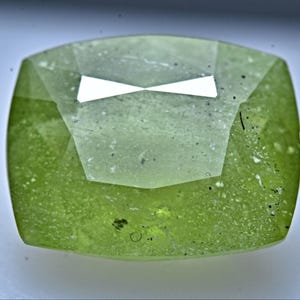 Pode incluir: Uma gema verde claro com um corte facetado e uma aparência ligeiramente nublada. A gema tem uma forma quadrada com cantos arredondados.